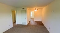 This single-floor space offers a functional and straightforward layout visible through the open living area. The front room features wall-to-wall carpet, providing a comfortable and practical surface underfoot, while the adjoining room transitions to hardwood flooring, creating a natural distinction between spaces. A ceiling fan with an integrated light fixture is centrally mounted, offering both air circulation and ambient lighting throughout. The white walls and standard-height ceilings contribute to a clean, neutral interior. A closet door is positioned centrally within the space, offering convenient storage access. Overhead HVAC vents and floor-level baseboard registers indicate a dual climate control system for year-round comfort. Natural light filters in through a window in the adjacent room, brightening the interior. A doorway to the left hints at additional rooms beyond the main living space. Overall, the layout is practical and open, with a straightforward design that provides a blank canvas adaptable to various furnishing and décor styles.