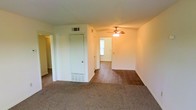This single-floor space offers a functional and straightforward layout visible through the open living area. The front room features wall-to-wall carpet, providing a comfortable and practical surface underfoot, while the adjoining room transitions to hardwood flooring, creating a natural distinction between spaces. A ceiling fan with an integrated light fixture is centrally mounted, offering both air circulation and ambient lighting throughout. The white walls and standard-height ceilings contribute to a clean, neutral interior. A closet door is positioned centrally within the space, offering convenient storage access. Overhead HVAC vents and floor-level baseboard registers indicate a dual climate control system for year-round comfort. Natural light filters in through a window in the adjacent room, brightening the interior. A doorway to the left hints at additional rooms beyond the main living space. Overall, the layout is practical and open, with a straightforward design that provides a blank canvas adaptable to various furnishing and décor styles.