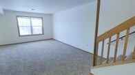 This multi-level residential space spans three floors, offering a well-structured layout designed for comfortable everyday living. The interior is characterized by a clean, neutral color palette, with white walls and light-toned carpeting that create a bright and open atmosphere throughout.

The living area pictured features plush wall-to-wall carpeting in a warm gray tone, providing a soft and practical underfoot surface. A window with wood-trimmed framing allows natural light to filter into the space, illuminating the room and contributing to its airy feel. The walls are smooth and white, offering a versatile backdrop suitable for a variety of furnishing styles.

A wooden staircase with light oak-toned balusters and handrails is prominently featured, connecting the floors of the home in a straightforward and functional design. The staircase adds a warm wooden accent that complements the neutral tones of the surrounding space.

The textured ceiling adds subtle visual depth to the rooms, while practical details such as baseboard trim and well-placed electrical outlets reflect a thoughtfully finished interior. Overall, the space presents a clean, move-in-ready environment across its three levels, well-suited for those seeking a functional and adaptable home layout.