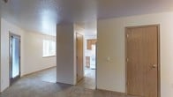 This single-floor residential unit offers a straightforward and functional layout designed for comfortable everyday living. The living area features wall-to-wall carpet and is illuminated by a window that allows natural light to fill the space, creating a bright and welcoming atmosphere. A textured ceiling runs throughout, adding a subtle visual element to the interior.

The space transitions seamlessly from the carpeted living area into a kitchen finished with tile flooring. The kitchen is equipped with light wood cabinetry providing ample storage, a white electric range, and a refrigerator, all arranged in a practical configuration suited for daily use. Under-cabinet space and open sightlines make the kitchen feel efficient and accessible.

Multiple interior doors are visible throughout the unit, suggesting the presence of additional rooms such as bedrooms, closets, or utility spaces that extend beyond the main living and kitchen areas. The neutral wall tones throughout create a clean, cohesive backdrop that is easy to personalize.

Overall, the unit presents a clean, move-in-ready interior with a sensible floor plan, a consistent neutral color palette, and a clear separation between the carpeted living zone and the tiled kitchen area, making it a practical and livable space.