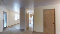 This single-floor residential unit offers a straightforward and functional layout designed for comfortable everyday living. The living area features wall-to-wall carpet and is illuminated by a window that allows natural light to fill the space, creating a bright and welcoming atmosphere. A textured ceiling runs throughout, adding a subtle visual element to the interior.

The space transitions seamlessly from the carpeted living area into a kitchen finished with tile flooring. The kitchen is equipped with light wood cabinetry providing ample storage, a white electric range, and a refrigerator, all arranged in a practical configuration suited for daily use. Under-cabinet space and open sightlines make the kitchen feel efficient and accessible.

Multiple interior doors are visible throughout the unit, suggesting the presence of additional rooms such as bedrooms, closets, or utility spaces that extend beyond the main living and kitchen areas. The neutral wall tones throughout create a clean, cohesive backdrop that is easy to personalize.

Overall, the unit presents a clean, move-in-ready interior with a sensible floor plan, a consistent neutral color palette, and a clear separation between the carpeted living zone and the tiled kitchen area, making it a practical and livable space.