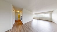 Located at 410 E Main St in Meriden, Connecticut, this one-bedroom, one-bathroom residence offers a well-organized single-floor layout with a practical and comfortable design. The living area is generously proportioned, featuring light-toned hardwood-style flooring and a wide bank of windows that invite abundant natural light and outdoor views of the surrounding greenery. A baseboard heating unit runs along the window wall, ensuring year-round comfort. The layout transitions seamlessly from the living space into a dedicated dining or flex area, also finished with matching flooring. The galley-style kitchen is efficiently designed with white cabinetry lining both sides, dark speckled countertops, a full suite of appliances including an electric range and refrigerator, and a window at the far end that keeps the space bright and airy. The bedroom is a comfortable size, carpeted for warmth, and features a wide sliding-door closet offering ample storage. A second closet is also available within the unit. The bathroom is finished with neutral tile work around the tub-shower combination, mosaic tile flooring, a white vanity, and a mirror. Multiple hallways connect the rooms, contributing to a clear and functional flow throughout the residence.
