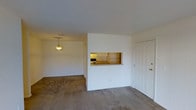 This single-floor residence offers a straightforward and functional layout designed for comfortable everyday living. The main living area is open and airy, featuring neutral wall-to-wall carpeting that extends throughout the space, contributing to a warm and quiet atmosphere. White walls and a smooth, painted ceiling keep the interior feeling bright and clean.

A standout architectural detail is the pass-through kitchen window cut into a central dividing wall, allowing a direct sightline into the kitchen area. This feature creates a sense of connectivity between the living and cooking spaces while maintaining a defined separation. The kitchen, visible through the pass-through, is equipped with cabinetry and counter space, providing efficient storage and workspace.

A pendant light fixture hangs in the transitional area between the living space and the kitchen zone, offering focused overhead lighting that adds a subtle decorative touch. A solid white entry door with brass hardware provides the main point of entry into the unit, positioned conveniently along one wall. Additional doorways lead to further areas of the home, suggesting additional rooms accessible from the main living space. Overall, the unit presents a clean, move-in ready environment with a practical open-concept sensibility.