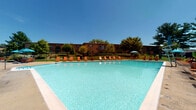 This single-story community pool facility is designed to serve residents with a range of outdoor and functional amenities. The centerpiece is a generously sized main swimming pool with clear, turquoise water, complemented by a separate wading pool for younger swimmers. The pool deck is lined with orange lounge chairs and includes a shaded umbrella area, all enclosed by a secure perimeter fence. Mature trees and well-kept green lawns surround the facility, with brick residential buildings visible in the background under open blue skies.

The interior support structure is constructed of concrete block masonry and features a staffed check-in counter where pool capacity is clearly posted — accommodating up to 83 in the main pool and 15 in the wading pool. The interior space includes a small utility area with basic appliances, a bulletin board with posted notices, and clearly marked gender-separated restroom facilities. Exposed wood beam ceilings and fluorescent lighting characterize the utilitarian interior. Access to the facility is provided through large metal doors that open to the surrounding landscaped grounds, offering views of green lawns and ornamental trees.