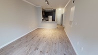This single-floor space showcases a thoughtfully designed open-concept layout that seamlessly connects the living area to the kitchen. Light wood-look flooring runs throughout, lending warmth and continuity to the interior. Crown molding lines the upper walls, adding a refined architectural detail to the otherwise clean, modern aesthetic.

The kitchen is outfitted with dark espresso cabinetry that creates a striking contrast against the light-toned surroundings. Stainless steel appliances, including a refrigerator and microwave, contribute a sleek, contemporary feel. A curved breakfast bar extends from the kitchen, offering a practical and stylish transition into the living space. Track lighting installed on the kitchen ceiling provides focused, adjustable illumination throughout the cooking and dining area.

The open living area benefits from the flow of natural and artificial light, making the space feel bright and airy. A hallway visible near the entryway suggests additional rooms or storage beyond the main living zone. Neutral wall tones throughout complement the flooring and cabinetry, resulting in a cohesive, move-in-ready interior well-suited for modern living.