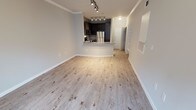 This single-floor space showcases a thoughtfully designed open-concept layout that seamlessly connects the living area to the kitchen. Light wood-look flooring runs throughout, lending warmth and continuity to the interior. Crown molding lines the upper walls, adding a refined architectural detail to the otherwise clean, modern aesthetic.

The kitchen is outfitted with dark espresso cabinetry that creates a striking contrast against the light-toned surroundings. Stainless steel appliances, including a refrigerator and microwave, contribute a sleek, contemporary feel. A curved breakfast bar extends from the kitchen, offering a practical and stylish transition into the living space. Track lighting installed on the kitchen ceiling provides focused, adjustable illumination throughout the cooking and dining area.

The open living area benefits from the flow of natural and artificial light, making the space feel bright and airy. A hallway visible near the entryway suggests additional rooms or storage beyond the main living zone. Neutral wall tones throughout complement the flooring and cabinetry, resulting in a cohesive, move-in-ready interior well-suited for modern living.