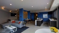 This contemporary single-floor space makes a strong visual impression through its use of bold cobalt blue accent walls contrasted against light gray tile flooring and a neutral ceiling with recessed lighting. The open-plan layout seamlessly connects multiple functional zones, creating a dynamic and inviting environment throughout.

The lounge area anchors one side of the space, furnished with a modern gray sofa accented by colorful throw pillows, a low round side table, and a wall-mounted flat-screen television. A patterned area rug grounds the seating arrangement and adds texture underfoot.

Adjacent to the lounge, a casual dining nook features a small table paired with sleek Scandinavian-style chairs, offering a comfortable spot for gathering or working. A decorative textured wall panel in warm tones serves as a striking focal point between the two zones, adding an artful architectural detail to the space.

On the opposite side, a long service counter with dark cabinetry and pendant lighting overhead provides a functional hub, ideal for a café-style service or reception area. The counter's clean lines and modern fixtures contribute to the overall polished aesthetic.

Pendant lights and recessed ceiling fixtures provide layered illumination throughout, while the open sightlines across the floor plan enhance the sense of spaciousness and flow.
