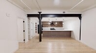 Located at 2309 First Ave N in Birmingham, Alabama, this single-floor unit masterfully combines industrial heritage with contemporary design. Light natural wood flooring runs throughout, complementing bright white walls and crisp trim. A defining architectural feature is the bold black steel structural column and exposed beam, lending an authentic industrial character to the open living area. The kitchen is a standout space, showcasing an expansive white quartz countertop island, taupe shaker-style cabinetry, black appliances, and a dramatic exposed brick accent wall adorned with rustic floating wood shelves and a sleek black range hood. Pendant lighting and an industrial-style flush mount fixture add warmth and definition to the kitchen zone. A dedicated utility area houses a stacked washer and dryer alongside generous built-in shelving set against another exposed brick wall. The bathroom features clean white tile flooring and a frameless glass walk-in shower with matte black hardware. Large black-framed sliding glass doors open directly to a private outdoor deck, filling the interior with natural light and offering a seamless indoor-outdoor connection. Frosted glass interior doors and louvered closet panels add refined detail throughout this well-appointed urban retreat.