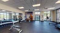 This single-floor fitness center offers a comprehensive and thoughtfully organized workout environment designed to accommodate a wide range of exercise needs. The space is divided into distinct functional zones, each tailored to a specific type of training.

The cardio area features a row of treadmills and elliptical machines positioned directly in front of expansive floor-to-ceiling windows, providing open views of lush outdoor greenery and allowing abundant natural light to fill the space. An overhead wall-mounted television adds convenience for users during longer sessions.

The strength training zone is equipped with cable machines, a Smith machine, and a full rack of dumbbells, all complemented by large wall mirrors that enhance the sense of space and support proper form. Dramatic large-scale murals depicting outdoor adventure scenes serve as bold visual focal points throughout the facility, adding an energizing and motivating atmosphere.

A dedicated free-motion and functional training area includes cycling bikes, medicine balls, and open floor space, finished with patterned carpet tiles that visually separate it from the rubber-floored strength and cardio sections. Ceiling fans and recessed panel lighting ensure consistent comfort and visibility throughout.

Additional amenities include a water fountain station and locker storage. The overall design is modern, clean, and purposefully laid out to support a complete fitness experience.