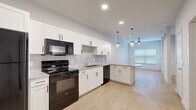 This efficiently designed single-floor unit offers a well-appointed layout that maximizes functionality across its compact footprint. The open kitchen serves as the centerpiece of the space, featuring crisp white shaker-style cabinetry with matte black hardware, granite-look countertops, and a tile backsplash. Black appliances — including a refrigerator, electric range, over-the-range microwave, and dishwasher — provide a bold contrast against the white cabinetry. A peninsula with pendant lighting overhead adds additional counter space and creates a natural division between the kitchen and the adjoining living area. Recessed lighting and a window allow natural light to brighten the open space. Light wood-look plank flooring runs throughout, lending warmth and visual continuity to the entire unit. The bathroom is well-equipped with a single vanity topped with granite-look counters, white shaker cabinetry, a full tub-shower combination with large-format tile surround, and matching light flooring. A dedicated closet provides convenient storage, and a separate laundry closet houses a stacked washer and dryer unit. The overall aesthetic is clean, modern, and move-in ready.