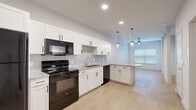 This efficiently designed single-floor unit offers a well-appointed layout that maximizes functionality across its compact footprint. The open kitchen serves as the centerpiece of the space, featuring crisp white shaker-style cabinetry with matte black hardware, granite-look countertops, and a tile backsplash. Black appliances — including a refrigerator, electric range, over-the-range microwave, and dishwasher — provide a bold contrast against the white cabinetry. A peninsula with pendant lighting overhead adds additional counter space and creates a natural division between the kitchen and the adjoining living area. Recessed lighting and a window allow natural light to brighten the open space. Light wood-look plank flooring runs throughout, lending warmth and visual continuity to the entire unit. The bathroom is well-equipped with a single vanity topped with granite-look counters, white shaker cabinetry, a full tub-shower combination with large-format tile surround, and matching light flooring. A dedicated closet provides convenient storage, and a separate laundry closet houses a stacked washer and dryer unit. The overall aesthetic is clean, modern, and move-in ready.