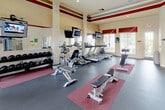 Located at Brandon Street in Woodstock, Georgia, this fitness center is a single-story community amenity space designed to support a full range of workout activities. The facility is characterized by notably high ceilings with crown molding and a bold burgundy and cream color scheme carried consistently throughout, lending the space a polished, upscale feel.

The main workout area is generously proportioned, accommodating a variety of cardio equipment including multiple treadmills and elliptical machines, each positioned in front of wall-mounted televisions for an engaging workout experience. The strength training zone features several cable and weight machines arranged on protective rubber flooring with red accent mats, along with free weights stored on a dedicated rack. Adjustable weight benches are distributed throughout for versatile training options.

Natural light enters through large windows and full-length glass doors that open directly to an outdoor pool area, providing pleasant outdoor views and a bright, airy atmosphere. A water fountain is conveniently mounted near the entrance. An adjoining room, visible through glass-paneled doors, appears to serve as a separate activity or community space with hardwood flooring. The combination of functional equipment, thoughtful layout, and refined architectural detailing makes this fitness center a standout community resource.