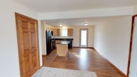 This single-floor space showcases an open-concept layout that seamlessly connects the kitchen and living areas, creating a spacious and functional environment. The kitchen is well-appointed with light maple cabinetry, dark granite-style countertops, and black appliances, including a refrigerator, microwave, and dishwasher. A central kitchen island with matching dark countertop provides additional workspace and serves as a natural dividing element between the kitchen and the adjoining living space. The flooring throughout the main area consists of rich, dark hardwood-style planks that add warmth and continuity. Natural light filters in through a window above the sink and a glass-paneled exterior door, brightening the interior and giving the space an open, airy feel. The walls are finished in a neutral cream tone that complements the warm wood accents found in the trim, doors, and cabinetry. A transition to carpet is visible at the edge of the living area, suggesting a defined zone for relaxation. Flush-mounted ceiling fixtures provide overhead lighting throughout the space.