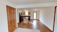 This single-floor space showcases an open-concept layout that seamlessly connects the kitchen and living areas, creating a spacious and functional environment. The kitchen is well-appointed with light maple cabinetry, dark granite-style countertops, and black appliances, including a refrigerator, microwave, and dishwasher. A central kitchen island with matching dark countertop provides additional workspace and serves as a natural dividing element between the kitchen and the adjoining living space. The flooring throughout the main area consists of rich, dark hardwood-style planks that add warmth and continuity. Natural light filters in through a window above the sink and a glass-paneled exterior door, brightening the interior and giving the space an open, airy feel. The walls are finished in a neutral cream tone that complements the warm wood accents found in the trim, doors, and cabinetry. A transition to carpet is visible at the edge of the living area, suggesting a defined zone for relaxation. Flush-mounted ceiling fixtures provide overhead lighting throughout the space.
