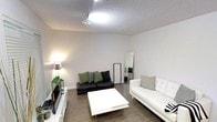 This single-floor space centers around a thoughtfully arranged living area that balances contrast and simplicity. The room features a notable ceiling height that contributes to an open, spacious atmosphere, complemented by a textured popcorn ceiling finish. Track lighting runs along the ceiling, providing directed illumination throughout the space.

The living area is anchored by two distinct seating pieces: a dark charcoal sofa adorned with green and black accent pillows, and a clean white sofa dressed with striped cushions. Between them sits a square white coffee table, reinforcing the room's black-and-white aesthetic. Dark hardwood-style flooring runs throughout, grounding the lighter elements of the space.

A floor lamp in the corner adds warm ambient lighting, while a flat-screen television is mounted on a low white media console along one wall. A tall full-length mirror leans against the wall, enhancing the sense of depth in the room. The neutral gray walls keep the space feeling calm and versatile. Overall, the layout is clean and functional, with a modern, minimalist character.