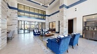 This impressive clubhouse delivers a sophisticated community experience across a generously sized single floor. The grand lobby immediately commands attention with its soaring double-height ceilings, bold blue architectural trim, and stacked-stone columns that lend a refined, contemporary character to the space. Large floor-to-ceiling windows flood the interior with natural light, while elegant tile flooring, statement chandeliers, and curated seating arrangements in deep navy blue create a polished yet welcoming atmosphere. A striking stone fireplace wall, complete with a mounted television and framed artwork, anchors one end of the main lounge area, providing a warm focal point. An adjoining lounge area features multiple seating groupings, a coffered tray ceiling, and views of the surrounding community through expansive windows. A built-in wet bar and dining area offer additional gathering space for residents. The game room is appointed with wood-look flooring, a coffered blue ceiling, a billiards table, ping pong, and direct access to an outdoor patio. The fitness facilities include two dedicated exercise rooms — one equipped with strength-training machines, free weights, and wall-to-wall mirrors, and a second dedicated to cardio with stationary bikes. A private office and connecting hallways complete this well-appointed clubhouse.
