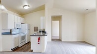 This single-floor residence is designed with a practical and cohesive layout that maximizes functionality throughout. The galley-style kitchen is lined on both sides with white cabinetry offering generous upper and lower storage, complemented by dark speckled countertops and wood-look vinyl flooring. White appliances — including an electric range, built-in microwave, refrigerator, and dishwasher — keep the space uniform and efficient. A peninsula with a double stainless steel sink extends toward the open living area, creating a natural transition between the kitchen and the carpeted main living space. The open-plan arrangement allows for flexible use of the living area, which connects seamlessly to additional rooms via a hallway. The bathroom features wood-look vinyl flooring, a single-sink vanity with matching dark countertop, a full tub and shower combination, and clean white fixtures throughout. A spacious walk-in closet fitted with wire shelving and hanging rods provides ample organization potential. A dedicated laundry area houses a stacked washer and dryer unit. Neutral cream-toned walls and consistent finishes throughout give the space a calm, move-in-ready ambiance.