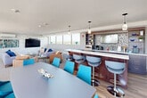 Located at 5220 Lakeshore Rd in Burlington, Ontario, this single-floor amenity lounge offers a thoughtfully designed open-plan layout that seamlessly blends multiple functional zones. The space is defined by expansive floor-to-ceiling windows and large glazed panels on multiple sides, flooding the interior with natural light and framing panoramic views of Lake Ontario and the surrounding Burlington skyline.

The lounge area features a textured stone accent wall serving as a striking focal point, complete with a wall-mounted flat-screen television and integrated climate control. Comfortable seating arrangements with neutral-toned sofas and blue accent cushions create a welcoming atmosphere. Adjacent to the lounge, a dedicated dining area accommodates a sizeable table with vibrant teal chairs, positioned directly beside the oversized windows to maximize the outdoor views.

A well-appointed bar and kitchenette runs along one side of the space, featuring rich dark wood cabinetry, a white quartz countertop, mosaic tile backsplash, open shelving, and modern appliances including a microwave and coffee station. Pendant lighting overhead adds a refined touch. Light wood-look flooring flows throughout, complementing the bright, contemporary palette. An outdoor terrace, accessible through large sliding doors, provides additional seating with direct lakefront views.