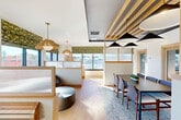 Located at 15 Pond Street Ct in Waltham, MA, this professionally designed workspace offers a well-appointed single-floor layout comprising five private offices, a dining room, and a connecting hallway. The open collaborative area is a standout feature, highlighted by an eye-catching ceiling with exposed wooden slat detailing, dramatic black pendant lights, and warm brass fixtures that create a sophisticated yet welcoming ambiance. Light hardwood flooring flows throughout the common areas, complementing the natural wood-framed glass partitions that divide spaces while maintaining an open, airy feel. Large windows dressed with patterned green Roman shades flood the space with natural light and offer views of the surrounding Waltham neighborhood. The dining and lounge area features a mix of seating options, including a built-in banquette, a conference-style table, and rounded lounge chairs, making it versatile for both formal and informal gatherings. Each private office is thoughtfully outfitted with built-in wooden desks, overhead shelving, brass task lighting, and textured carpeting, providing a focused and comfortable environment for individual work. A larger office with a glass whiteboard and collaborative seating adds flexibility for small meetings. The hallway connects all spaces seamlessly, with warm-toned wood doors and frosted glass panels maintaining privacy throughout.