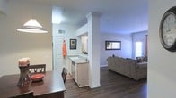 This single-story residence offers a thoughtfully arranged floor plan that balances comfort and functionality across a generous layout. The open-concept design connects the kitchen, dining, and living areas into one cohesive social space, fostering a seamless flow ideal for everyday living and entertaining. The kitchen is framed by a decorative column with crown molding detail and features white cabinetry, a sink with dishwasher, and a layout that opens directly to the dining area. Rich, dark hardwood-style flooring extends throughout the main living spaces, adding warmth and a sense of continuity. Crown molding accents the ceilings, lending a polished architectural touch throughout. The living room is bright and welcoming, benefiting from natural light filtering through large windows. The floor plan, as seen from above, reveals multiple bedrooms — including a spacious primary bedroom — each offering ample room for furnishings. A dedicated bathroom and additional washroom areas are conveniently accessible. The overall layout is well-organized with distinct zones for sleeping, living, and dining, making this a practical and comfortable single-floor home suited for a variety of lifestyles.