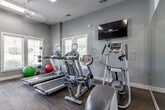 The Poplar Glen Apartment Homes fitness center is a thoughtfully designed, single-floor exercise facility built to support a well-rounded workout routine. The space is finished with cool-toned blue-gray walls, durable mixed flooring of hardwood and carpet, and ceiling fans that help maintain air circulation throughout. Large windows on multiple walls flood the room with natural light and offer views of the surrounding greenery, creating an inviting and energizing atmosphere.

The cardio zone is well-stocked with multiple treadmills equipped with individual digital displays, elliptical trainers, a stationary bike, and a stair climber, catering to a range of cardio preferences. A wall-mounted flat-screen TV is positioned for easy viewing during workouts. The strength and flexibility area includes a rowing machine, a dumbbell rack with a full range of weights, medicine balls, and stability balls, providing options for both resistance training and functional fitness.

Full-length mirrors line one wall, amplifying the sense of space and allowing users to monitor their form. The facility is accessed through a glass-paneled door and features a dedicated entrance, a water station, and ample room to move freely between equipment. The overall layout is efficient and user-friendly, making this fitness center a comprehensive amenity for residents.
