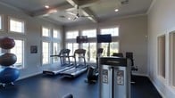 This single-story fitness center is designed to provide a bright, functional workout space. The room is defined by its coffered ceiling with a large ceiling fan, which enhances air circulation and adds an architectural touch to the space. Recessed lighting complements the natural light that floods in through numerous large windows along the walls, offering views of the surrounding greenery and creating an open, energizing atmosphere.

The exercise floor is finished with a durable dark rubberized surface, practical for high-traffic fitness use. The space is outfitted with Life Fitness branded cardio equipment, including multiple treadmills and elliptical-style machines, positioned to allow users to enjoy the outdoor views while working out. Two wall-mounted flat-screen televisions are strategically placed for easy viewing from the cardio equipment.

The neutral gray walls and white-trimmed windows contribute to a clean, contemporary aesthetic throughout the room. The overall ambiance is airy and motivating, combining well-considered design details with practical fitness amenities to create an inviting environment for exercise.
