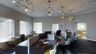 This contemporary workspace offers a well-designed, single-floor environment built for productivity and collaboration. The room is anchored by a series of striking geometric cube-framed pendant lights with exposed Edison-style bulbs, lending an industrial-modern aesthetic that sets the tone for the entire space. Neutral gray walls provide a calm, focused backdrop, while large windows dressed with horizontal blinds allow natural light to filter in and brighten the interior. The workstation area features curved, dark wood-toned desks arranged in a thoughtful layout, each equipped with desktop computers and separated by low-profile dividers that offer a degree of privacy without sacrificing the open feel of the room. Modern white and yellow accent chairs add a pop of color and contemporary flair to the seating throughout. A cozy lounge nook with patterned upholstered chairs is visible near the windows, providing an informal area for casual meetings or relaxed work. The overall ambiance is clean, polished, and energizing — a functional yet stylish environment that balances professionalism with modern design sensibility, making it well-suited for business centers, study lounges, or shared office environments.