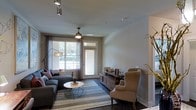 This single-floor space offers a thoughtfully designed interior that balances comfort and contemporary style. The living area serves as the centerpiece of the space, showcasing dark hardwood flooring that grounds the room with warmth and elegance. A large window and a glass-paneled door flood the space with natural light, framing views of the greenery outside while providing direct access to an outdoor area. Sheer curtains complement the horizontal blinds, offering flexible light control throughout the day. The ceiling features a classic ceiling fan with a built-in light fixture, while track lighting overhead adds a modern, functional touch. The living room is furnished with a plush sectional sofa accented with decorative pillows, paired with a round white coffee table resting atop a striped area rug. A wooden media console serves as a focal point along one wall, flanked by a comfortable wingback accent chair in a warm neutral tone. Decorative branches add an organic, artistic element to the space. The clean white walls and neutral color palette create a serene, inviting ambiance that is both modern and livable, making this a highly functional and aesthetically cohesive living environment.