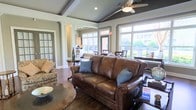 This single-floor space exudes a warm and inviting atmosphere, thoughtfully designed to welcome guests with an upscale yet comfortable aesthetic. The main lounge area is anchored by a rich brown leather sofa accented with blue throw pillows, complemented by traditional patterned seating and dark wood accent tables that add a classic touch to the décor.

Architectural details elevate the space considerably. Tray ceilings with a deep gray-blue finish are framed by crisp white crown molding and decorative ceiling beams, creating a refined overhead focal point. A ceiling fan with a light fixture provides both functionality and style. Recessed lighting adds a warm, layered glow throughout.

Expansive multi-pane windows line the perimeter, flooding the interior with natural light and offering views of the surrounding greenery and exterior environment. The durable hardwood flooring runs throughout, grounding the space with warmth and continuity.

Beyond the lounge, the layout opens to additional areas, including what appears to be a kitchen or refreshment station with cabinetry visible in the background, as well as a dining area with dark wood chairs. Columns and glass-paneled doors define transitions between spaces while maintaining an open, connected feel. Overall, the space balances elegance and practicality in a well-appointed, multi-functional layout.