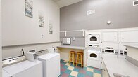 This shared laundry facility is a thoughtfully designed, single-level space that maximizes functionality within a compact footprint. The room features notably high ceilings that lend an open, airy feel, complemented by crisp white walls accented by a gray feature wall housing the primary appliance bank. Multiple top-loading washing machines and stacked dryer units provide ample laundry capacity, while a dedicated utility sink with cabinetry below offers additional convenience for hand-washing or pre-treating garments. A built-in folding counter runs along one wall, paired with two wooden bar stools—creating a practical workspace for sorting and folding. A wall-mounted shelf above the counter provides additional storage and hang-dry space. Natural light filters into the space through black-framed windows, offering views to an outdoor courtyard area. The distinctive multi-colored checkered vinyl flooring in teal, mauve, and cream adds a lively, welcoming character to the room. Decorative wall art and small accent pieces contribute a homey touch. The space is well-ventilated with ceiling-mounted HVAC vents, and includes a payment/information terminal and safety equipment for user convenience.