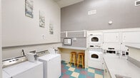 This shared laundry facility is a thoughtfully designed, single-level space that maximizes functionality within a compact footprint. The room features notably high ceilings that lend an open, airy feel, complemented by crisp white walls accented by a gray feature wall housing the primary appliance bank. Multiple top-loading washing machines and stacked dryer units provide ample laundry capacity, while a dedicated utility sink with cabinetry below offers additional convenience for hand-washing or pre-treating garments. A built-in folding counter runs along one wall, paired with two wooden bar stools—creating a practical workspace for sorting and folding. A wall-mounted shelf above the counter provides additional storage and hang-dry space. Natural light filters into the space through black-framed windows, offering views to an outdoor courtyard area. The distinctive multi-colored checkered vinyl flooring in teal, mauve, and cream adds a lively, welcoming character to the room. Decorative wall art and small accent pieces contribute a homey touch. The space is well-ventilated with ceiling-mounted HVAC vents, and includes a payment/information terminal and safety equipment for user convenience.