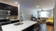 This single-floor residence offers a thoughtfully designed open-concept layout that seamlessly connects the kitchen, dining, and living areas, creating a cohesive and functional living environment. The kitchen serves as the centerpiece, featuring a large white quartz island with an undermount sink and stylish faucet, paired with rich dark cabinetry that extends to the ceiling for ample storage. Recessed lighting illuminates the space effectively throughout. The open dining area accommodates a round table with upholstered chairs, positioned conveniently between the kitchen and living room. The living area is anchored by a ceiling fan, large windows dressed with floor-length draping curtains that allow natural light to filter in, and a media console with a wall-mounted television. A gallery-style arrangement of framed artwork adorns the main wall, adding a polished, curated aesthetic to the shared living space. Dark hardwood flooring flows consistently throughout, grounding the neutral, light-gray color palette. A private bedroom is accessible directly from the main living area, visible through an interior doorway and featuring coordinated furnishings. The overall ambiance is modern, clean, and inviting, with a balanced mix of dark and light tones that lend the space a sophisticated yet comfortable character.