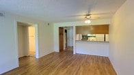 This single-floor space offers a straightforward, functional layout designed for comfortable everyday living. The main living area is open and airy, featuring warm hardwood-style flooring that extends throughout the space, creating a cohesive and inviting atmosphere. A ceiling fan with a light fixture is centrally positioned, providing both airflow and ambient lighting to the room.

The kitchen is seamlessly integrated into the living space via a half-wall peninsula, which features a stone-look countertop and warm wood-toned cabinetry overhead. The kitchen is equipped with white appliances, including a refrigerator, stove, and microwave, set against teal-accented cabinetry that adds a subtle pop of color. The open-plan design ensures the kitchen remains connected to the living area, making it well-suited for both daily routines and casual entertaining.

Adjacent to the main living space, a hallway leads to additional rooms, with wood cabinetry and open shelving visible, offering generous storage solutions. The textured popcorn ceiling is consistent throughout, and the neutral white walls provide a clean, versatile backdrop. Overall, the space presents a practical and well-connected layout with warm finishes and ample storage.