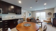 This thoughtfully designed single-floor residence offers a cohesive open-concept layout that seamlessly connects the kitchen, dining, and living areas. The kitchen is outfitted with rich dark cabinetry, granite countertops, stainless steel appliances including a full-size refrigerator, electric range, and built-in microwave, complemented by wood-style flooring that extends into the dining area. A round dining table positioned adjacent to the kitchen provides a functional and inviting space for meals. The living room features plush carpeting, recessed lighting, and a large sliding glass door that opens to a private balcony, filling the space with natural light and offering open exterior views. Two well-proportioned bedrooms provide comfortable retreats, each with soft carpeting, recessed lighting, and natural light from windows. Both bathrooms are finished with granite-topped vanities, dark cabinetry, wood-style flooring, and tiled soaking tub and shower combinations. Neutral wall tones and consistent finishes throughout the residence create a clean, cohesive aesthetic. The dollhouse view confirms an efficient and well-organized floor plan that maximizes livability within a compact, single-level footprint.