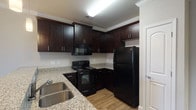 The Autumn floor plan is a well-appointed 1-bedroom, 1-bathroom unit spread across a single level, offering a thoughtfully designed layout that balances functionality and comfort. The open-concept living area features warm wood-look flooring and crown molding, creating an inviting atmosphere with natural light flowing in through a glass exterior door. The kitchen is a standout feature, showcasing rich dark espresso cabinetry, granite countertops, a double stainless steel sink, and a full suite of black appliances including a refrigerator, electric range, and over-the-range microwave. A peninsula with pendant lighting connects the kitchen to the living space, making it ideal for everyday living and entertaining. The bedroom is generously sized, outfitted with plush carpet, a ceiling fan, and a large window that allows ample natural light and outdoor views. The bathroom features tile flooring, a dark vanity with a granite countertop, a large mirror with vanity lighting, and a full shower-tub combo. Throughout the unit, neutral tones and consistent finishes contribute to a cohesive and modern aesthetic.