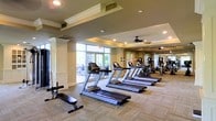 This fitness center offers a spacious, thoughtfully designed workout environment that balances functionality with refined aesthetics. The facility features a generous open floor plan finished with neutral-toned carpeting and crisp white decorative columns that lend a polished, upscale feel to the space.

A row of treadmills forms the centerpiece of the cardio zone, complemented by a variety of additional cardio and strength-training machines positioned throughout the room. Ceiling fans are mounted above to promote air circulation during workouts, while recessed lighting provides even, comfortable illumination throughout.

Large floor-to-ceiling windows along one wall flood the interior with abundant natural light and offer views of the outdoor surroundings, creating an inviting and energizing atmosphere. The tray ceiling detailing with crown molding adds an architectural sophistication seldom found in fitness facilities.

A mirrored wall on one side of the room visually expands the space while allowing users to monitor their form during exercise. The overall ambiance is clean, bright, and well-maintained, reflecting a high standard of upkeep. This fitness center is well-suited for a broad range of workout routines and provides an encouraging environment for users of all fitness levels.