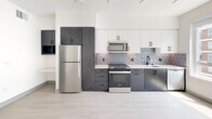 This thoughtfully designed single-floor unit offers a clean, modern aesthetic carried throughout every room. Light wood-look flooring runs seamlessly across the open-plan living area, creating a bright and airy atmosphere complemented by a ceiling fan overhead. The kitchen is a standout feature, showcasing two-tone cabinetry with charcoal lower cabinets and crisp white uppers, paired with a geometric patterned tile backsplash. Stainless steel appliances — including a refrigerator, gas range, and over-the-range microwave — and a matte black faucet at the sink add a contemporary edge. The bathroom is finished with large-format light stone-look tile flooring, a white vanity with matte black hardware, wall-mounted cabinetry, and a tub-shower combination accented with wood-tone tile surround. A dedicated walk-in closet provides generous storage with wire shelving lining the walls. Rounding out the unit's convenience is an in-unit laundry area featuring a stacked Whirlpool washer and dryer. The consistent use of neutral tones, clean lines, and modern fixtures throughout creates a cohesive and functional living space.