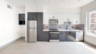 This thoughtfully designed single-floor unit offers a clean, modern aesthetic carried throughout every room. Light wood-look flooring runs seamlessly across the open-plan living area, creating a bright and airy atmosphere complemented by a ceiling fan overhead. The kitchen is a standout feature, showcasing two-tone cabinetry with charcoal lower cabinets and crisp white uppers, paired with a geometric patterned tile backsplash. Stainless steel appliances — including a refrigerator, gas range, and over-the-range microwave — and a matte black faucet at the sink add a contemporary edge. The bathroom is finished with large-format light stone-look tile flooring, a white vanity with matte black hardware, wall-mounted cabinetry, and a tub-shower combination accented with wood-tone tile surround. A dedicated walk-in closet provides generous storage with wire shelving lining the walls. Rounding out the unit's convenience is an in-unit laundry area featuring a stacked Whirlpool washer and dryer. The consistent use of neutral tones, clean lines, and modern fixtures throughout creates a cohesive and functional living space.