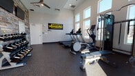 This fitness center is designed to support a comprehensive workout experience within a single open floor layout. The space features soaring ceilings that create an airy, open atmosphere, complemented by large, floor-to-ceiling windows that flood the room with natural light and offer views of the surrounding exterior. The flooring throughout is durable rubber gym matting, practical for both heavy equipment and functional training.

The cardio section is outfitted with multiple treadmills and elliptical machines positioned to take advantage of the natural light from the large windows. A cable resistance machine and adjustable weight bench round out the strength training options. A full dumbbell rack lines one wall, offering a wide range of free weights for varied strength workouts.

A dedicated functional training area features a well-stocked equipment rack holding medicine balls, stability balls, resistance bands, and yoga mats — catering to flexibility, balance, and conditioning routines. A mounted flat-screen television is positioned on the accent wall for entertainment during workouts.

The design incorporates a striking stacked-stone accent wall that adds a warm, textural contrast to the otherwise clean, modern interior. A ceiling fan aids in air circulation, and a motivational banner reading "Don't Be Average, Be Awesome" adds an energizing touch to the overall ambiance.