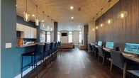Located at 3995 S. Hamilton Rd. in Groveport, Ohio, this single-floor community clubhouse is designed with a contemporary, open-concept layout that seamlessly blends social, recreational, and functional spaces. Rich dark hardwood flooring runs throughout, anchoring a cohesive and upscale aesthetic.

The kitchen and bar area serves as a central hub, featuring white shaker cabinetry, bold teal accent walls, a stainless steel refrigerator, and an extended bar counter lined with sleek black leather high-back stools. A dramatic cluster of glass pendant lights hangs overhead, adding warmth and visual interest to the space.

Adjacent to the kitchen, a dedicated workstation area provides a row of individual desks with ample surface space, ideal for productivity. Nearby, a lounge area offers a relaxed retreat centered around a full-height stacked stone fireplace, complemented by comfortable seating, patterned area rugs, and large windows that invite natural light and outdoor views.

The game area, boldly branded with a "GAME ON" sign set against a reclaimed wood accent wall, features a billiards table and casual seating, creating a lively entertainment zone. The overall ambiance of the space balances modern design with inviting warmth, making it a well-rounded gathering destination for the community.