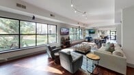 Located at 191–199 Carolina Point Pkwy in Greenville, SC, this single-floor community clubhouse is designed to offer residents a welcoming and versatile gathering space. The layout flows openly across several distinct functional zones, all united by rich hardwood-style flooring and a clean, contemporary aesthetic.

The main lounge area is expansive and bathed in natural light, courtesy of oversized black-framed windows that frame views of the surrounding greenery. Track lighting overhead complements the natural illumination, while multiple seating arrangements — including plush sofas, accent chairs, and glass-topped coffee tables — create an inviting atmosphere suited for both relaxation and socializing. Wall-mounted flat-screen televisions are positioned throughout for added convenience.

A secondary lounge zone provides an equally comfortable setting with tray ceiling detailing, additional large windows, and curated furniture groupings that encourage conversation.

The business center stands out with vibrant, colorful abstract artwork displayed gallery-style along one wall, complementing the functional wooden desks, ergonomic seating, and computer workstations available for resident use.

The demonstration kitchen is well-appointed with gray shaker-style cabinetry, a white quartz island with bar seating, stainless steel appliances including a double oven and refrigerator, a subway tile backsplash, and pendant lighting — all reflecting a polished, modern design sensibility. A framed mirror in the adjacent hallway adds depth and visual interest to the space.