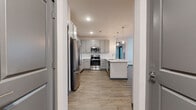Located in Nokomis, Florida, this single-floor residence offers a thoughtfully designed open-concept layout with contemporary finishes throughout. The kitchen serves as the heart of the home, featuring grey shaker-style cabinetry with brushed nickel hardware, crisp white quartz countertops, a white tile backsplash, and a full suite of stainless steel appliances including a range, microwave, and refrigerator. A peninsula island with pendant globe lighting overhead provides additional prep space and naturally connects the kitchen to the living area. The living space is generously proportioned, bathed in natural light through large windows and a glass-paneled entry door, and finished with warm wood-look luxury vinyl plank flooring. Ceiling fans are present in both the living and bedroom areas for year-round comfort. The bedroom features plush carpeting, a window with blinds for natural light and privacy, and a ceiling fan. The bathroom is well-appointed with grey cabinetry, a white quartz vanity top, wood-look flooring, and a full tub-shower combination finished with large-format grey tile surround and a built-in shelf niche. The cohesive use of neutral tones and modern fixtures throughout creates a clean, inviting atmosphere.