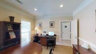 This single-floor office space offers a clean, professional environment designed for productivity and functionality. The main work area is anchored by a dark wood desk equipped with a computer, printer, and office essentials, complemented by ergonomic black chairs. Warm, neutral-toned walls paired with crown molding give the space a polished, traditional aesthetic. Recessed ceiling lights provide even, ambient illumination throughout, while a large window dressed with horizontal blinds invites natural light and creates an open, airy atmosphere. Framed artwork adorns the walls, adding a refined touch to the professional setting. A round wooden table near the entrance offers an additional surface for meetings or collaborative work. The space transitions seamlessly into a tiled hallway leading to a dedicated restroom, ensuring convenience for occupants and visitors alike. The combination of carpeted work areas and tiled transitional spaces reflects a thoughtful approach to both comfort and practicality. Overall, this office space balances a welcoming ambiance with the functional amenities needed for an efficient and organized work environment.