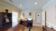 This single-floor office space offers a clean, professional environment designed for productivity and functionality. The main work area is anchored by a dark wood desk equipped with a computer, printer, and office essentials, complemented by ergonomic black chairs. Warm, neutral-toned walls paired with crown molding give the space a polished, traditional aesthetic. Recessed ceiling lights provide even, ambient illumination throughout, while a large window dressed with horizontal blinds invites natural light and creates an open, airy atmosphere. Framed artwork adorns the walls, adding a refined touch to the professional setting. A round wooden table near the entrance offers an additional surface for meetings or collaborative work. The space transitions seamlessly into a tiled hallway leading to a dedicated restroom, ensuring convenience for occupants and visitors alike. The combination of carpeted work areas and tiled transitional spaces reflects a thoughtful approach to both comfort and practicality. Overall, this office space balances a welcoming ambiance with the functional amenities needed for an efficient and organized work environment.