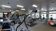 This fitness center is a single-floor facility thoughtfully designed to accommodate a comprehensive workout routine. The space is outfitted with a diverse selection of exercise equipment, including elliptical machines, treadmills, and weight benches, providing options for both cardiovascular and strength training. Protective rubber floor mats are positioned beneath equipment to ensure safety and durability.

Floor-to-ceiling mirrors line the walls, creating a sense of expanded space while allowing users to monitor their form during exercise. A ceiling-mounted flat-screen television offers entertainment and keeps members informed during workouts. Overhead fluorescent lighting provides consistent, bright illumination throughout the room, ensuring a well-lit environment at all hours.

Ventilation is supported by a ceiling fan, contributing to a comfortable workout atmosphere. Large windows along one wall invite natural light into the space and offer views of the surrounding outdoor environment, adding an open and airy feel to the interior. The neutral-toned walls and durable carpeted flooring complete the functional and practical design of this fitness facility, making it a well-rounded space suited for users of all fitness levels.