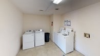 This shared laundry room is a practical, single-floor facility designed to meet the everyday laundry needs of its users. The space is laid out efficiently, housing multiple top-loading washing machines and full-sized dryers arranged along the walls for easy access. The room features light-toned walls and tile flooring, creating a clean and functional environment. Overhead fluorescent lighting ensures the space is well-lit throughout. A built-in folding counter extends along one wall, providing a dedicated surface for sorting and folding laundry. Washer hookup connections are recessed into the wall, reflecting a tidy and organized installation. Safety is addressed with a fire extinguisher mounted and accessible. Informational signage is posted on the walls, offering guidance on machine usage and laundry services. A waste bin is conveniently placed within the room. The space connects through a paneled door to an adjacent area, adding to the overall accessibility of the facility. Overall, this laundry room is a well-maintained, straightforward utility space designed with practicality and user convenience in mind.