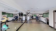 This contemporary fitness center is thoughtfully designed to support a full range of workout routines across a single, open floor. The space is divided into distinct functional zones, with a dedicated cardio area and a separate strength training section, allowing for efficient use of the floor plan.

The cardio zone features a row of high-tech treadmills and elliptical machines, each equipped with individual screens for entertainment and workout tracking. Exposed black ductwork and piping run along the ceiling in this area, lending an industrial edge to the design, while circular halo pendant lights provide warm, focused illumination throughout.

The strength training area is outfitted with a comprehensive selection of cable machines and resistance equipment arranged to maximize space and accessibility. Floor-to-ceiling mirrors line the walls, visually expanding the room and allowing users to monitor their form during exercises. Accent walls in a bold green add a vibrant, energetic feel to the environment.

The flooring throughout is a durable, cushioned green rubber surface with dark accent striping, designed for both comfort and safety. The overall aesthetic is clean and modern, with white walls and ceilings contrasting the darker equipment and industrial ceiling details, creating an inviting and motivating environment for fitness enthusiasts.