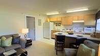 This single-floor residence features an open-concept layout that seamlessly integrates the kitchen and living areas into one cohesive space. The kitchen is outfitted with light maple cabinetry offering ample storage, white appliances including a full-size refrigerator and electric range, and a double-basin sink positioned beneath under-cabinet lighting. Fluorescent overhead lighting illuminates the workspace efficiently. A peninsula counter extends from the kitchen, providing a casual dining or entertaining space accommodated by dark-framed bar stools with upholstered seats. The counter surface is light-colored and clean, lending a bright, open feel to the area. The flooring transitions from tile in the kitchen zone to plush neutral-toned carpeting in the living area, creating a subtle visual distinction between functional and relaxing spaces. The living area is warmly lit by a table lamp and opens toward a doorway leading to an adjacent room. The walls are finished in soft, warm neutral tones throughout, contributing to a calm and cohesive ambiance. The overall layout is practical and efficiently organized, making good use of the available footprint.