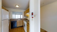 This single-floor residence at Amber Gardens offers a practical and well-organized layout. The kitchen is a standout feature, equipped with light wood cabinetry that provides ample storage space, dark granite-style countertops, and a full suite of appliances including a white gas range and a black dishwasher. An under-cabinet range hood sits above the stove, adding both functionality and a clean, streamlined look. The kitchen also includes a double basin sink positioned beneath a multi-pane window, allowing natural light to filter in and brighten the workspace. The warm hardwood-style flooring throughout the kitchen complements the cabinetry and contributes to a cohesive aesthetic. A structural column near the kitchen entry adds architectural definition while subtly separating the kitchen from the adjoining carpeted living area, which is partially visible from this vantage point. Safety features such as a mounted fire extinguisher and smoke detector are also present. The overall ambiance is practical and inviting, with a neutral color palette that provides a versatile backdrop for a variety of interior styles.