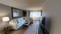 This single-floor space centers around a welcoming living room designed with a clean, neutral aesthetic. The room is anchored by a generously sized white sectional sofa, complemented by coordinating accent pillows and a dark-stained wood coffee table that adds contrast to the light carpeted flooring. A large flat-screen television is positioned opposite the seating area, making the space well-suited for both relaxing and entertaining.

Natural light filters into the room through a wide window dressed with vertical blinds, creating a bright and airy atmosphere. A pair of floor and side-table lamps provide warm supplemental lighting. A tall indoor plant adds a touch of greenery and visual height to one corner of the room.

A large framed map-style artwork adorns the accent wall, lending character and a point of visual interest to the space. The textured ceiling and neutral wall tones contribute to the room's understated, comfortable ambiance. Overall, the living area is thoughtfully arranged to maximize both function and comfort within a cohesive, modern-casual design scheme.
